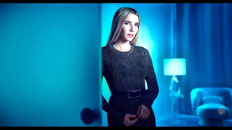 "American Horror Story": Emma Roberts in a dark, textured dress stands by a door in a softly lit, blue-toned room. A lamp and armchair are visible, creating a serene ambiance.