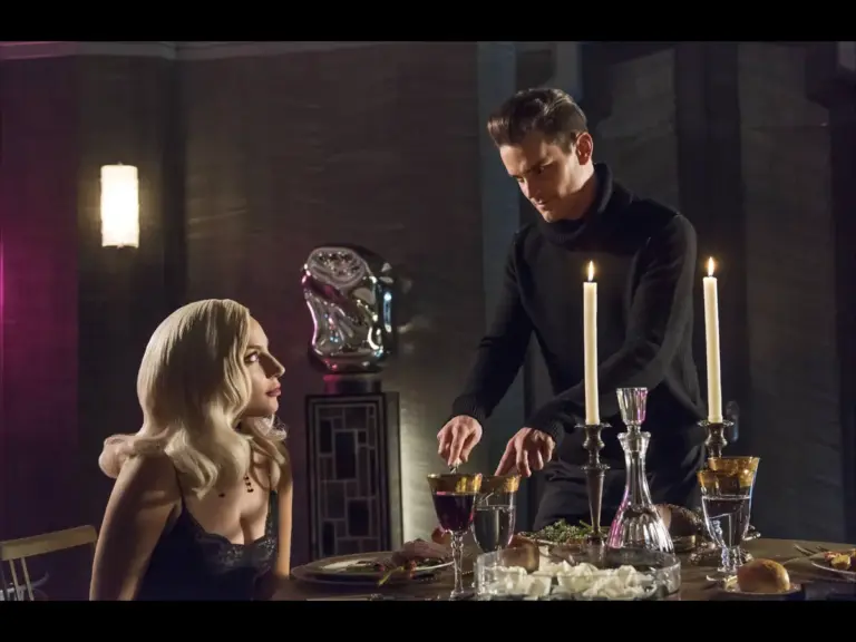 "American Horror Story" : A woman with blonde hair in black attire sits at a candlelit dinner table, looking at a man in black who is serving food, creating a mysterious mood.