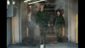 Rebecca Hall, Dan Stevens, and Kaylee Hottle in Godzilla X Kong: The New Empire walk confidently through a smoky corridor. Two wear green tactical gear, while one wears a casual outfit with sunglasses. The mood is intense.