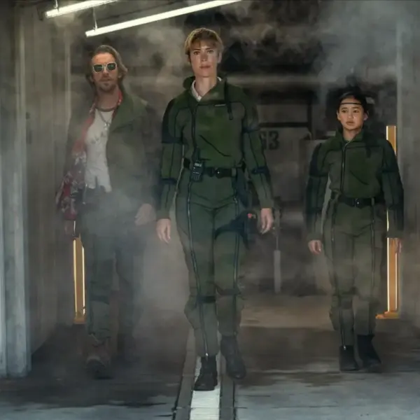Rebecca Hall, Dan Stevens, and Kaylee Hottle in Godzilla X Kong: The New Empire walk confidently through a smoky corridor. Two wear green tactical gear, while one wears a casual outfit with sunglasses. The mood is intense.