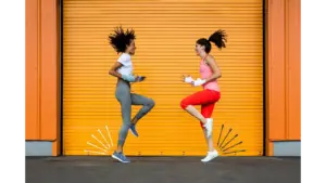 Two healthy women in athletic wear jump playfully in front of a bright orange corrugated metal garage door.