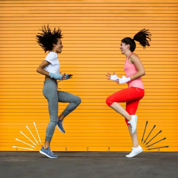 Two healthy women in athletic wear jump playfully in front of a bright orange corrugated metal garage door.