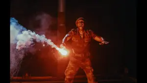 Mahershala Ali in Jurassic World: Rebirth (2025) A man in camouflage attire holds a firework, preparing to light it against a dark background.
