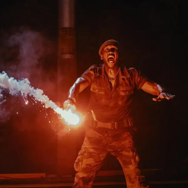 Mahershala Ali in Jurassic World: Rebirth (2025) A man in camouflage attire holds a firework, preparing to light it against a dark background.