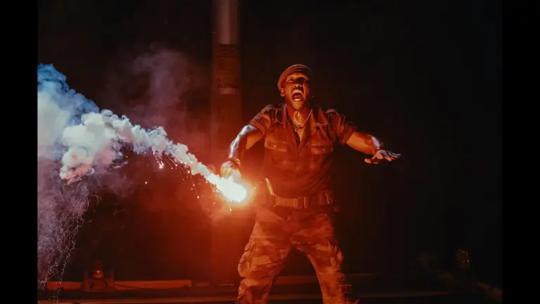 Mahershala Ali in Jurassic World: Rebirth (2025) A man in camouflage attire holds a firework, preparing to light it against a dark background.