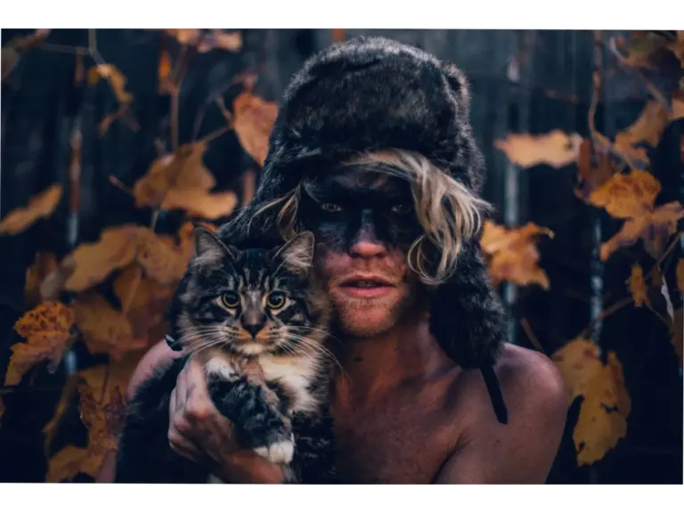 A man wearing a fur hat and dark face paint holds a tabby cat, both staring intensely — a moody pet lookalikes portrait.