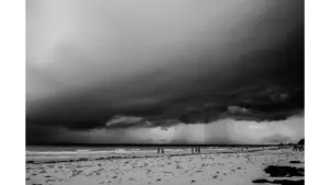 Monochrome view of an impending storm over a beach with people walking along the shore, capturing the dramatic mood of storm travel.