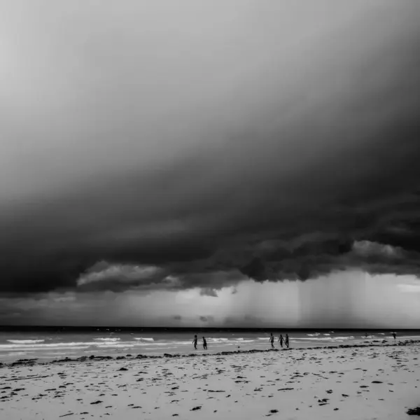 Monochrome view of an impending storm over a beach with people walking along the shore, capturing the dramatic mood of storm travel.