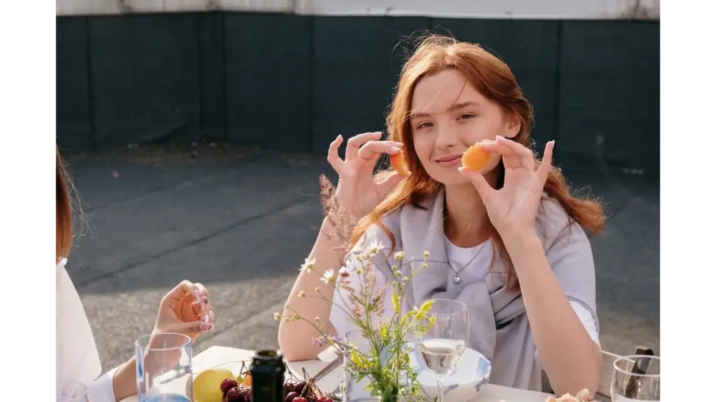 Smiling young woman enjoys a casual brunch outdoors, holding snacks and surrounded by friends, capturing the carefree vibe of a girl dinner moment.