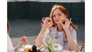Smiling young woman enjoys a casual brunch outdoors, holding snacks and surrounded by friends, capturing the carefree vibe of a girl dinner moment.