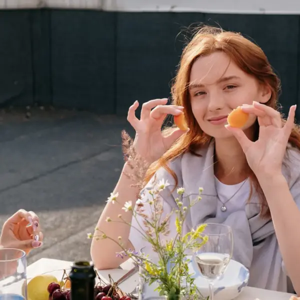 Smiling young woman enjoys a casual brunch outdoors, holding snacks and surrounded by friends, capturing the carefree vibe of a girl dinner moment.
