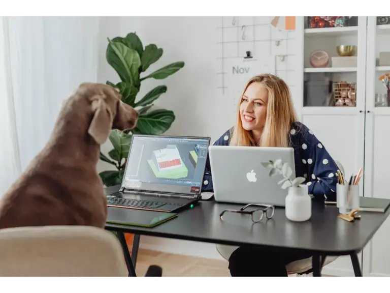 A woman smiles at her dog while working on laptops at her home office desk, a perfect snapshot of how Call Preference shifts when real life and remote work blend together.