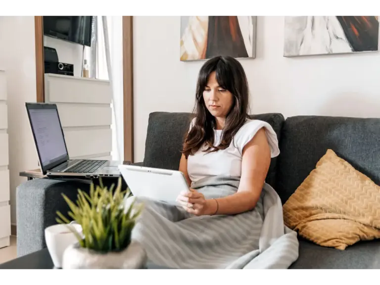 Woman on sofa with tablet and laptop, enjoying a cozy home work environment that perfectly fits the energy of the remote boom.