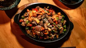 Delicious grilled steak bites with peppers and tomatoes in a hot skillet on wooden table