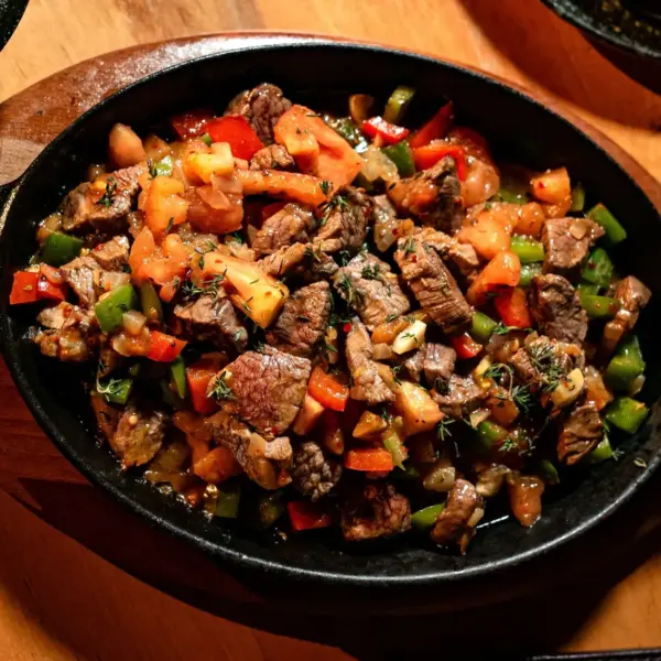 Delicious grilled steak bites with peppers and tomatoes in a hot skillet on wooden table