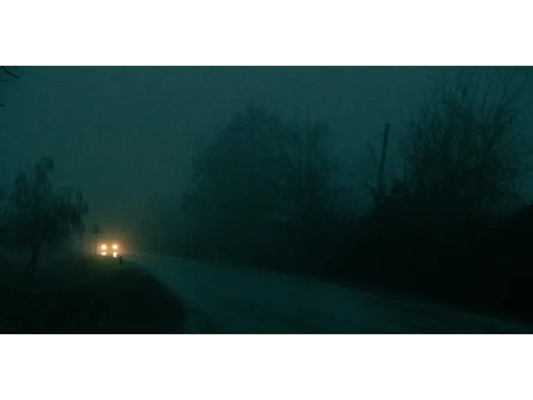 A car's headlights pierce through dense fog on a dark, eerie haunted road at night