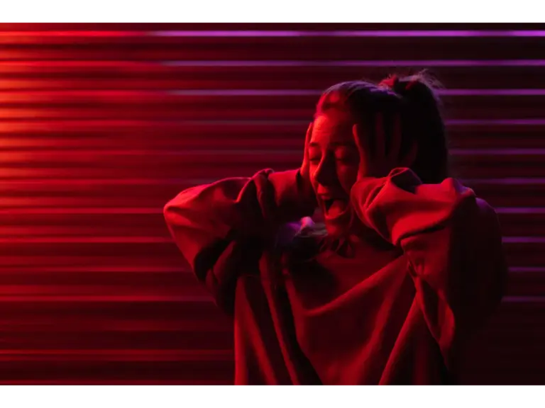 Woman with hands on head, expressing stress in neon red lighting, a sharp visual echo of the horror origins that make tension feel cinematic.