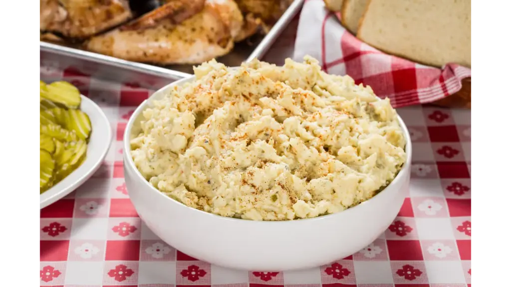 Creamy viral potato salad in a white bowl with BBQ and pickles on a checkered tablecloth