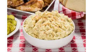 Creamy viral potato salad in a white bowl with BBQ and pickles on a checkered tablecloth