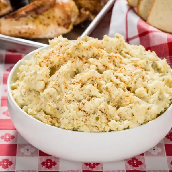 Creamy viral potato salad in a white bowl with BBQ and pickles on a checkered tablecloth