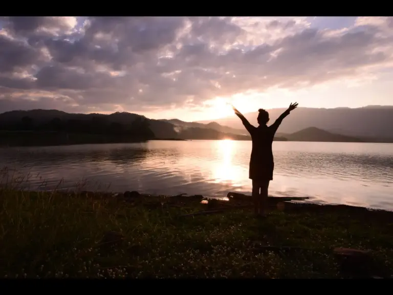 In daily devotioal, a woman lifts her arms as she basks in the goodness of God near a lake.