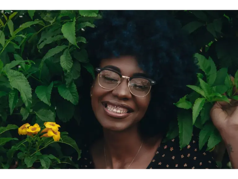 A cheerful woman with afro hair grins amidst lush green leaves and yellow flowers outdoors, beautifully highlighting natural frames that complement her vibrant style.