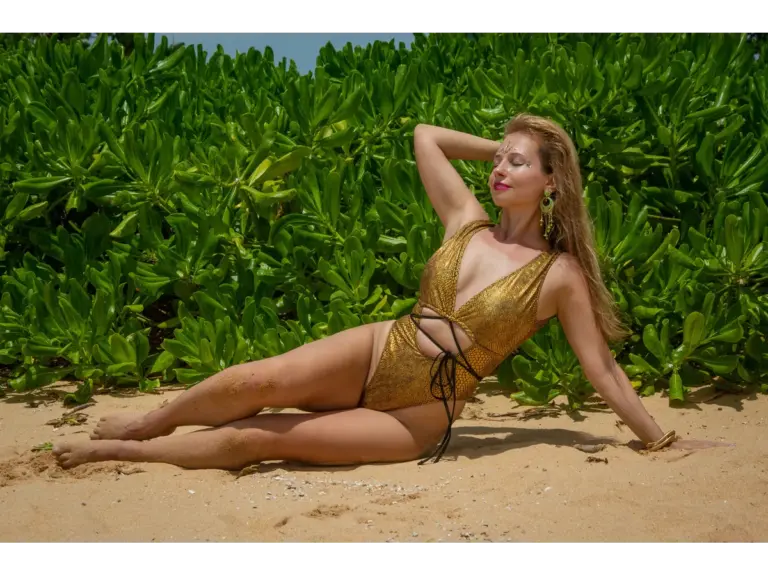 Elegant woman in a gold swimsuit lounges on a sunny beach, shadowed by lush green plants, capturing the warm, effortless energy of this year’s Swimwear Trends.