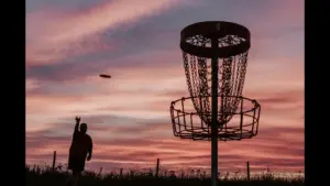 A silhouette of a person throwing a frisbee towards a disc golf basket at sunset, with colorful clouds in the sky.