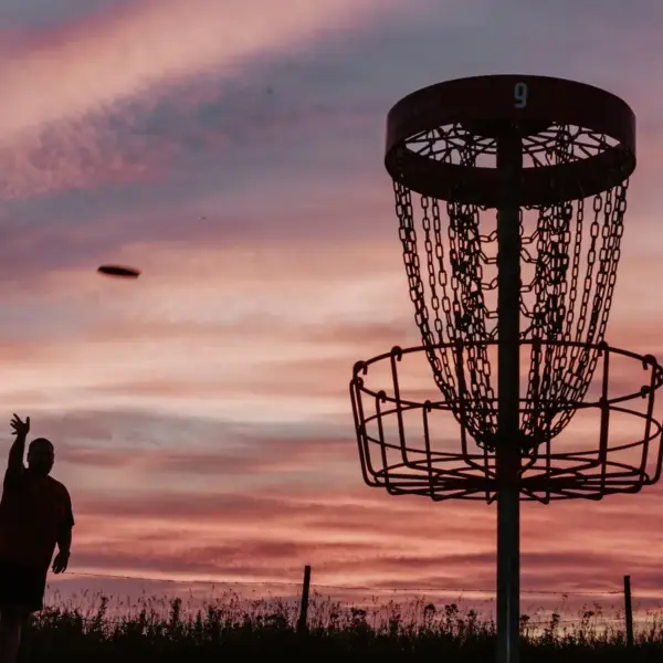 A silhouette of a person throwing a frisbee towards a disc golf basket at sunset, with colorful clouds in the sky.