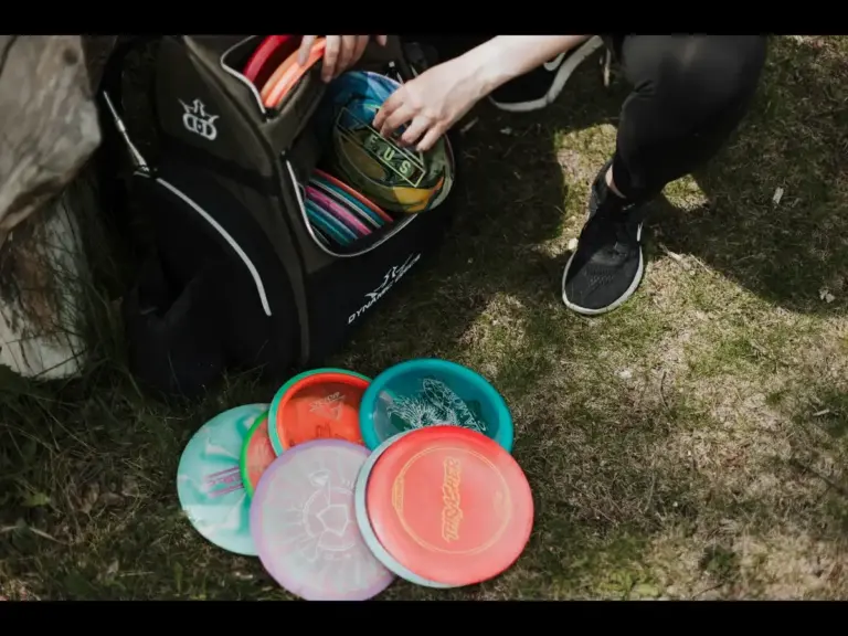 A person crouches next to a disc golf bag, reaching for discs inside, with several colorful discs laid out on the ground.