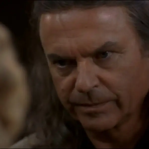 Sam Neill as Merlin in "Merlin's Apprentice" tv series staring intently, conveying determination. His expression is serious, suggesting a tense or dramatic situation. Blurred figure in foreground.