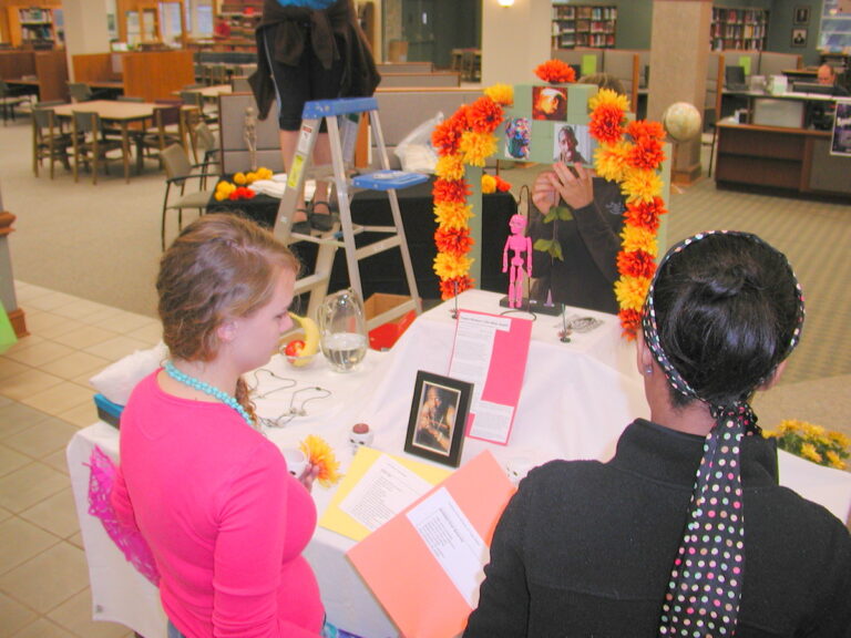 Two people observe a colorful library display decorated with flowers, photos, and papers honoring Tupac Legacy, symbolizing cultural remembrance and creativity.