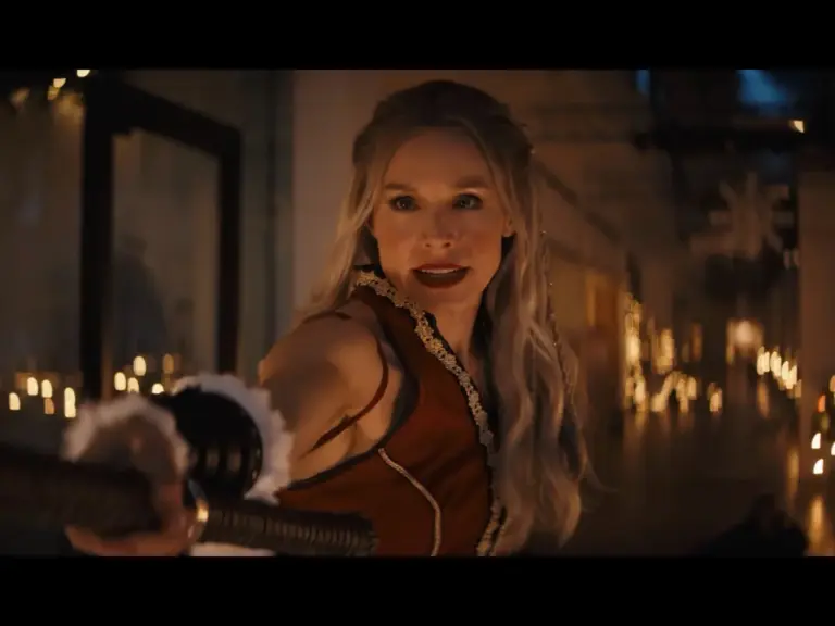 Kristen Bell as Mrs. Claus with long blonde hair, wearing a red dress, striking a confident pose while holding a weapon, set in a dimly lit environment adorned with candles.