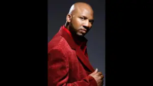 Samuel Monroe Jr, actor from "Menace II Society," wearing a red velvet suit, striking a confident pose against a dark background.