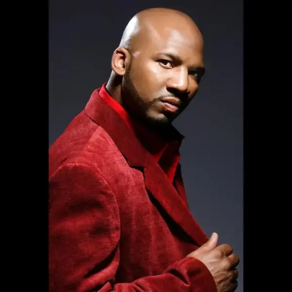 Samuel Monroe Jr, actor from "Menace II Society," wearing a red velvet suit, striking a confident pose against a dark background.