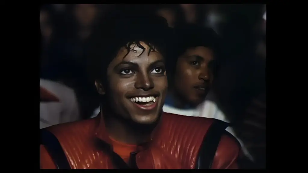 Music video for "Thriller" featuring Michael Jackson: a smiling man with curly hair wearing a red jacket, sitting in a crowd.