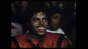 Music video for "Thriller" featuring Michael Jackson: a smiling man with curly hair wearing a red jacket, sitting in a crowd.