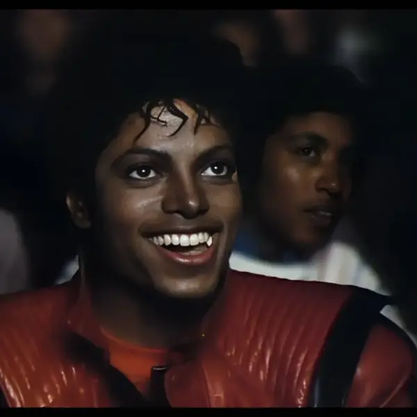 Music video for "Thriller" featuring Michael Jackson: a smiling man with curly hair wearing a red jacket, sitting in a crowd.