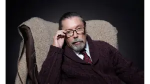 Tim Curry, wearing glasses and a maroon cardigan, sits in a cozy chair, looking thoughtfully at the camera against a dark background.