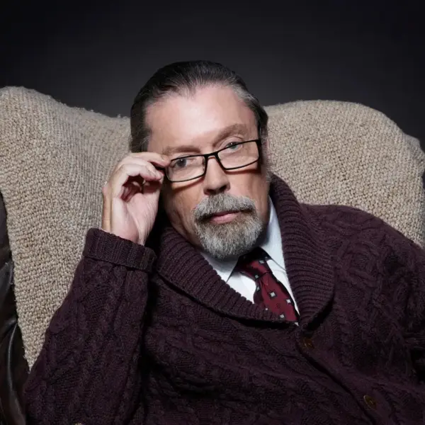 Tim Curry, wearing glasses and a maroon cardigan, sits in a cozy chair, looking thoughtfully at the camera against a dark background.