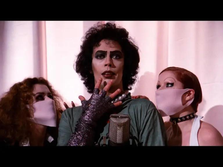 A scene from The Rocky Horror Picture Show (1975) featuring Tim Curry in dramatic makeup and costume, with two other individuals wearing masks in the background.