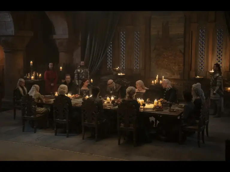A large group of people seated around a long dining table in a dimly lit, medieval-style room, with candles providing light. Some individuals are dressed in elaborate costumes, while others are in simpler attire. The atmosphere appears formal and serious.