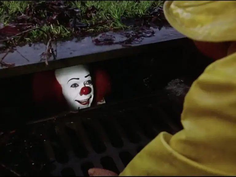 Tim Curry in IT (1990) appears from a storm drain while a person in a yellow raincoat looks on.