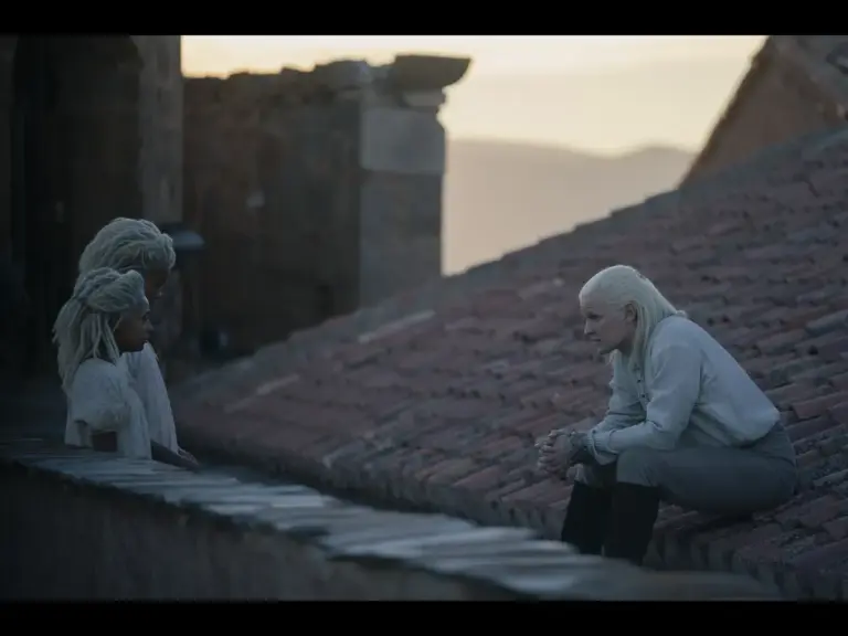 Two figures with white hair sitting on a rooftop, engaged in a conversation with another figure with a braided hairstyle. The background features a scenic view and a soft, warm sunset.