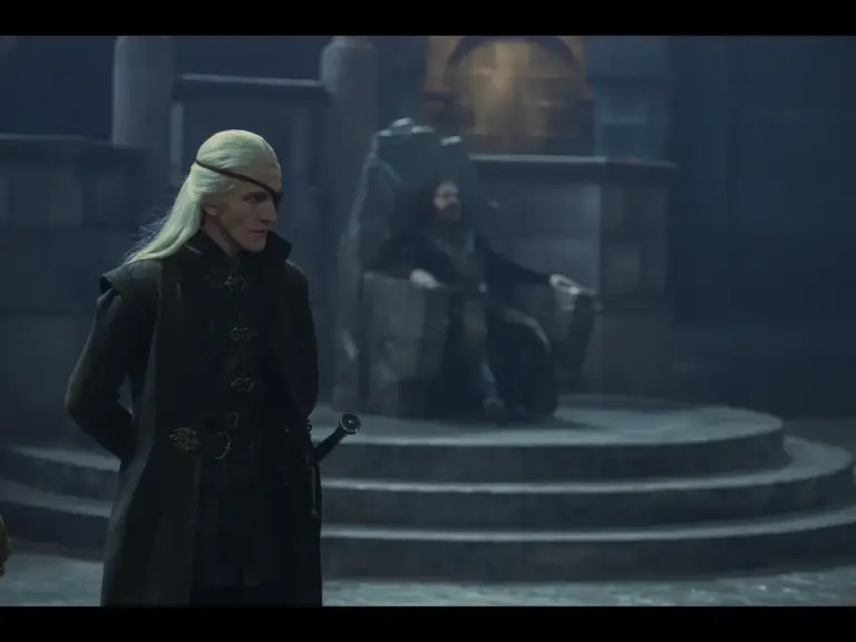 A scene from a fantasy series featuring two characters, one standing with long white hair and wearing dark clothing, and another seated in a stone throne in the background.