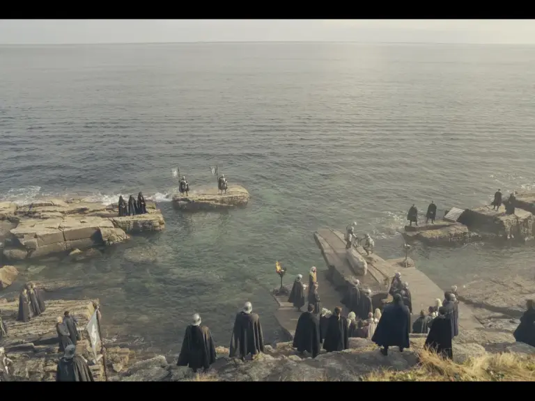 A group of figures in dark cloaks standing near rocky shores, with some near the water and others on a dock. The scene is set against a calm sea in the background.