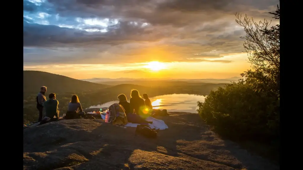 A group of people watching the sunset from a rocky viewpoint, overlooking a lake and mountains in the distance, with a dramatic sky.