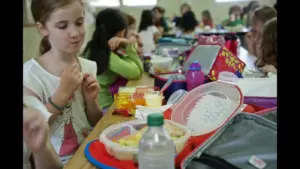 A group of children sitting at a table eating lunch, with various lunchboxes and food containers visible. A girl in the foreground is eating and looking at her food. school lunch, packed lunch, back-to-school