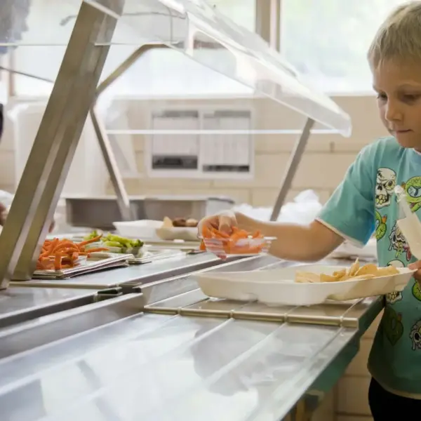 A young boy in a blue t-shirt serving food at a cafeteria line. School cafeteria prices, school lunch, back to school