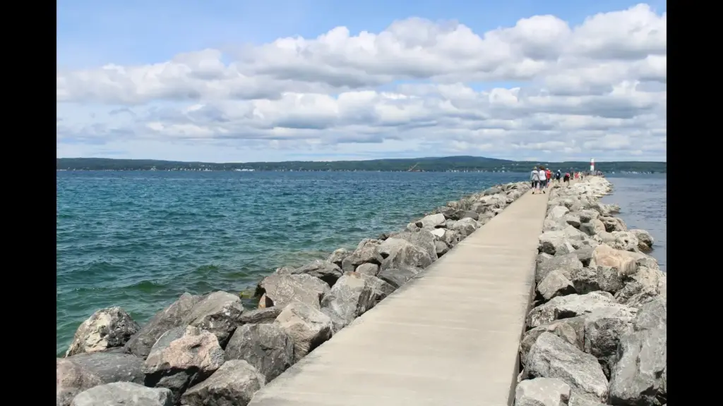 A rocky pier extending into a blue body of water, with cloudy skies above and people walking along the path. Traverse City, MI, USA